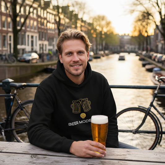 Beer Mode On premium black hoodie lifestyle photo Amsterdam canal beer garden Netherlands - Dutch man early 30s blonde hair outdoor picnic table canal houses bicycles Dutch pilsner golden hour - bold white text graphic craft beer enthusiast European beer culture Amsterdam lifestyle Dutch fashion canal city unisex hoodie Guapo Originals Premium Designer Collection €39.99 Spain